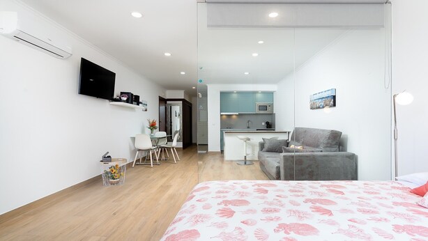 A modern hotel room with a bed, a desk, a chair, a sofa, a kitchenette, and a TV.