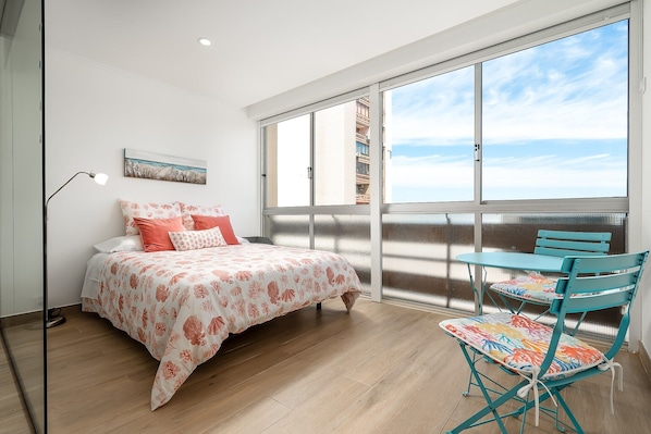 A bedroom with a bed, a table, and chairs, a large window, and a painting on the wall.
