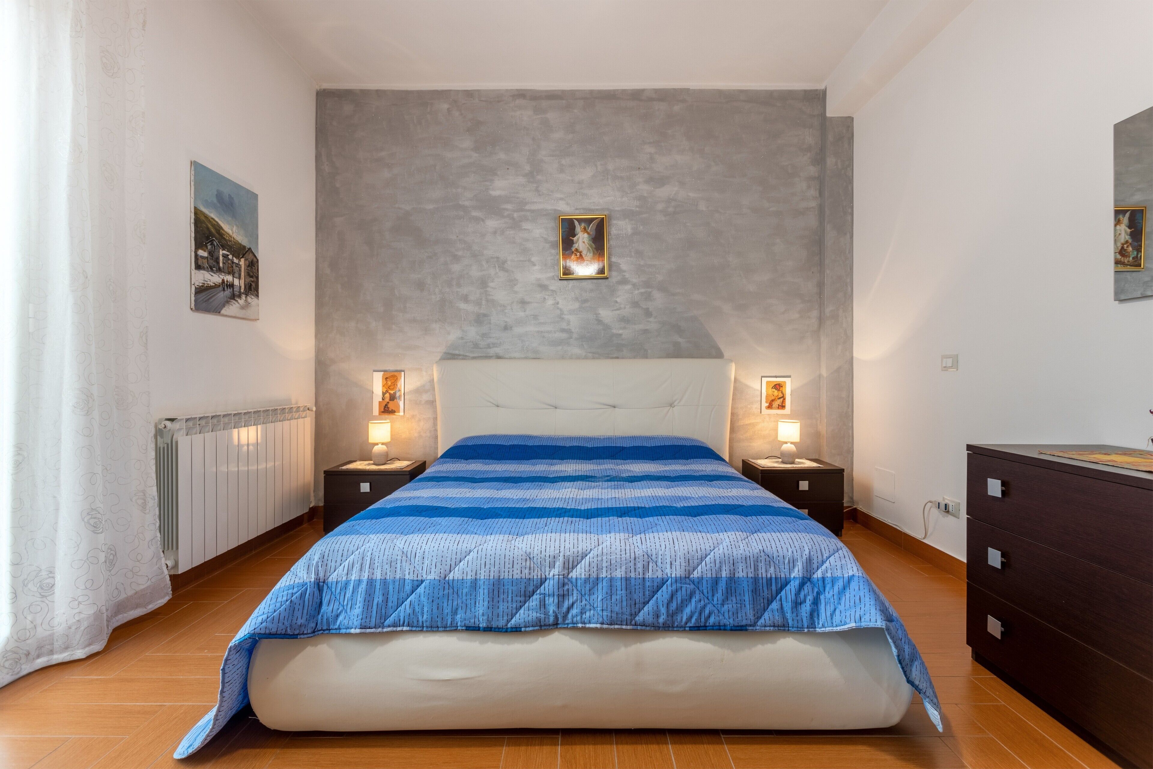 A bedroom with a large bed, two bedside tables with lamps, a dresser, and a radiator.