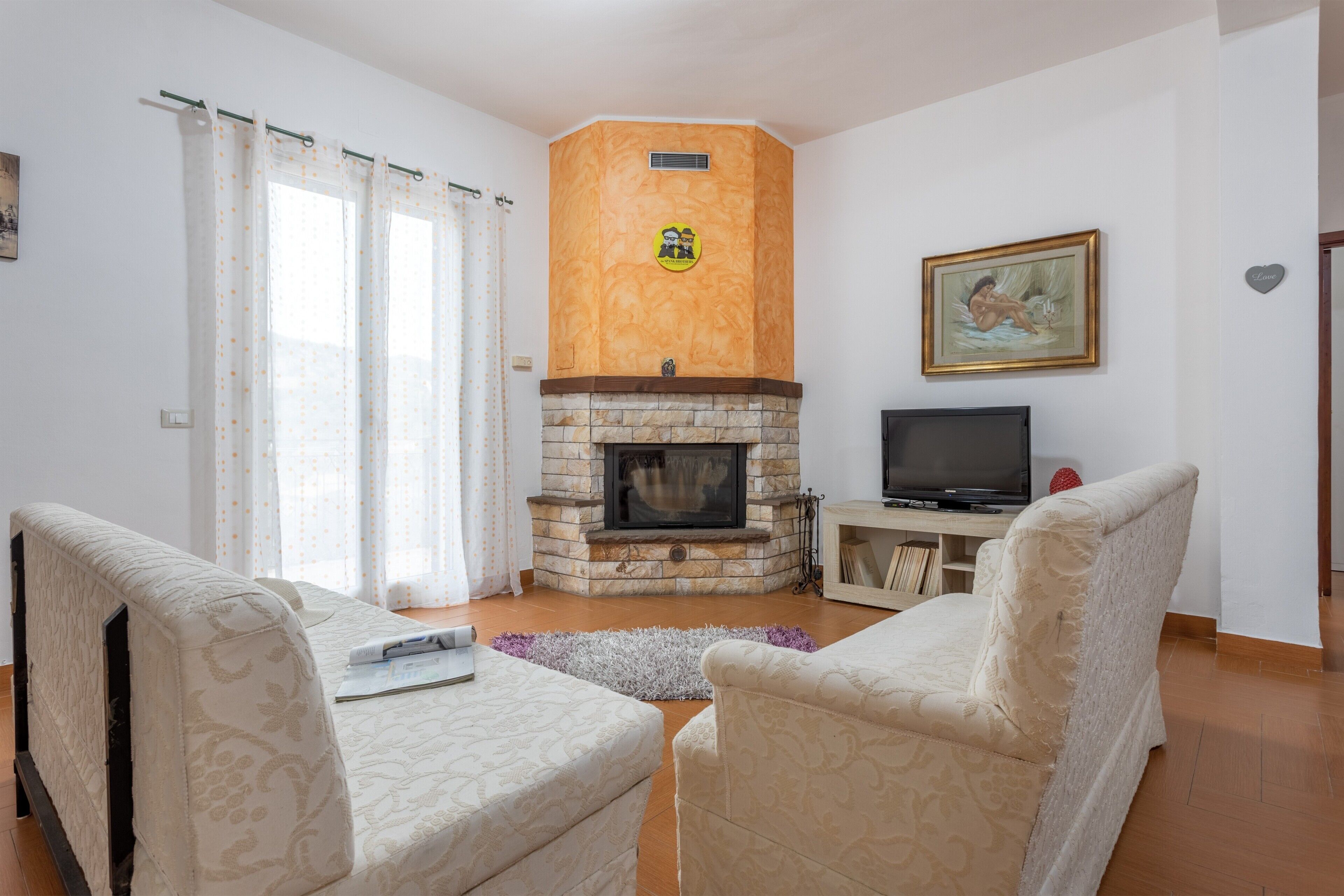 Un salon avec une cheminée, deux fauteuils, un téléviseur et un tableau accroché au mur.