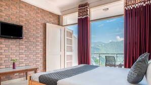 Une chambre d’hôtel avec un mur en briques, un téléviseur à écran plat, un lit en bois, un balcon avec des rideaux rouges et une vue sur la montagne.