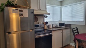 A compact kitchen with a stainless steel refrigerator, a microwave, an oven, and a sink. There is a calendar on the refrigerator door, and a window with blinds in the background. - Main Floor Suite (Stettler)
