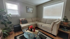 A living room with a beige sectional sofa, a glass coffee table with decorative items, a black ottoman, and a large potted plant. - Main Floor Suite (Stettler)