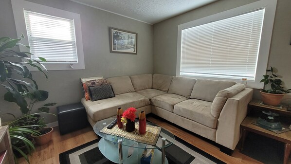 A living room with a beige sectional sofa, a glass coffee table with decorative items, a black ottoman, and a large potted plant. - Main Floor Suite (Stettler)