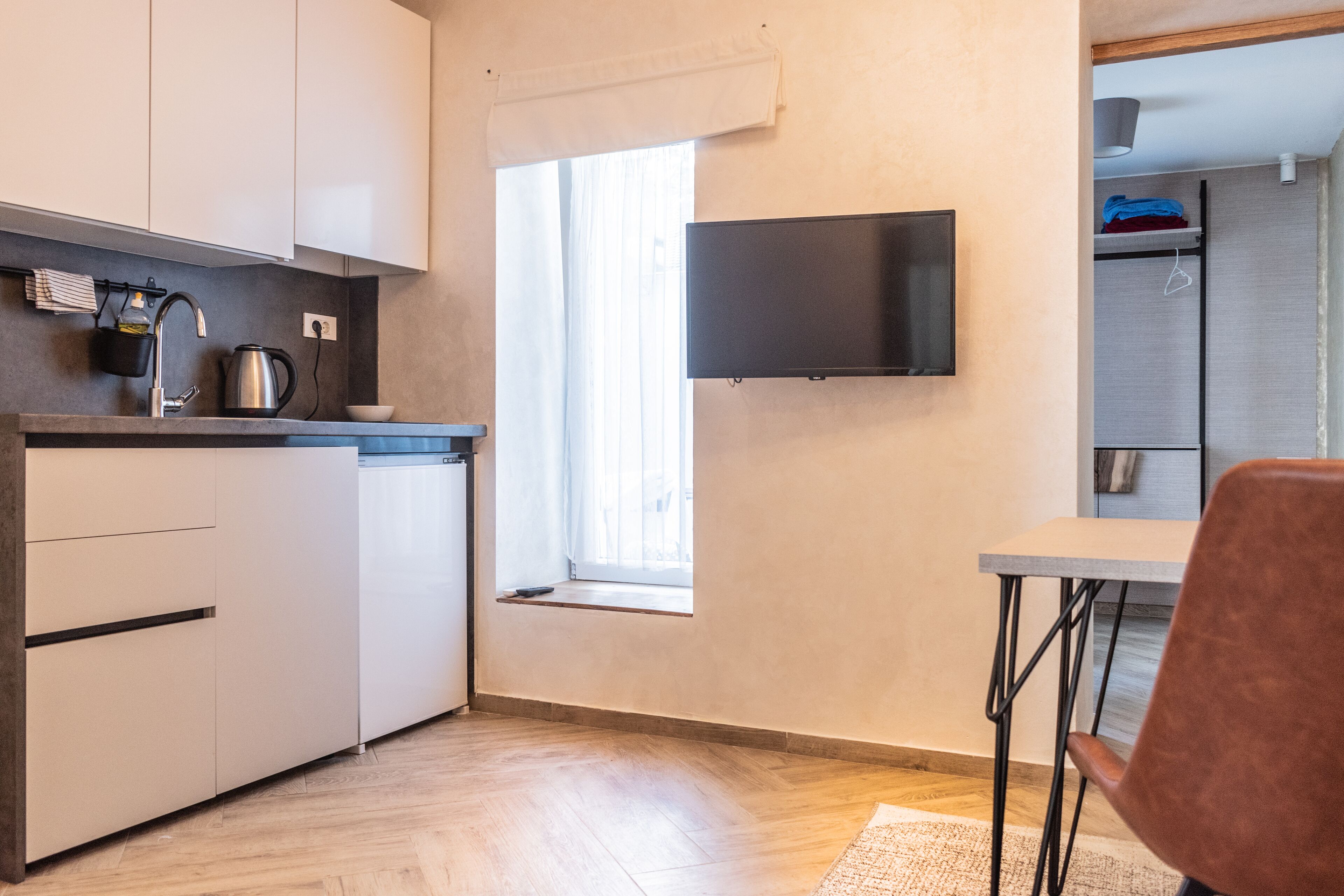 A compact kitchen with a flat-screen TV, a window, and a dining area with a chair.