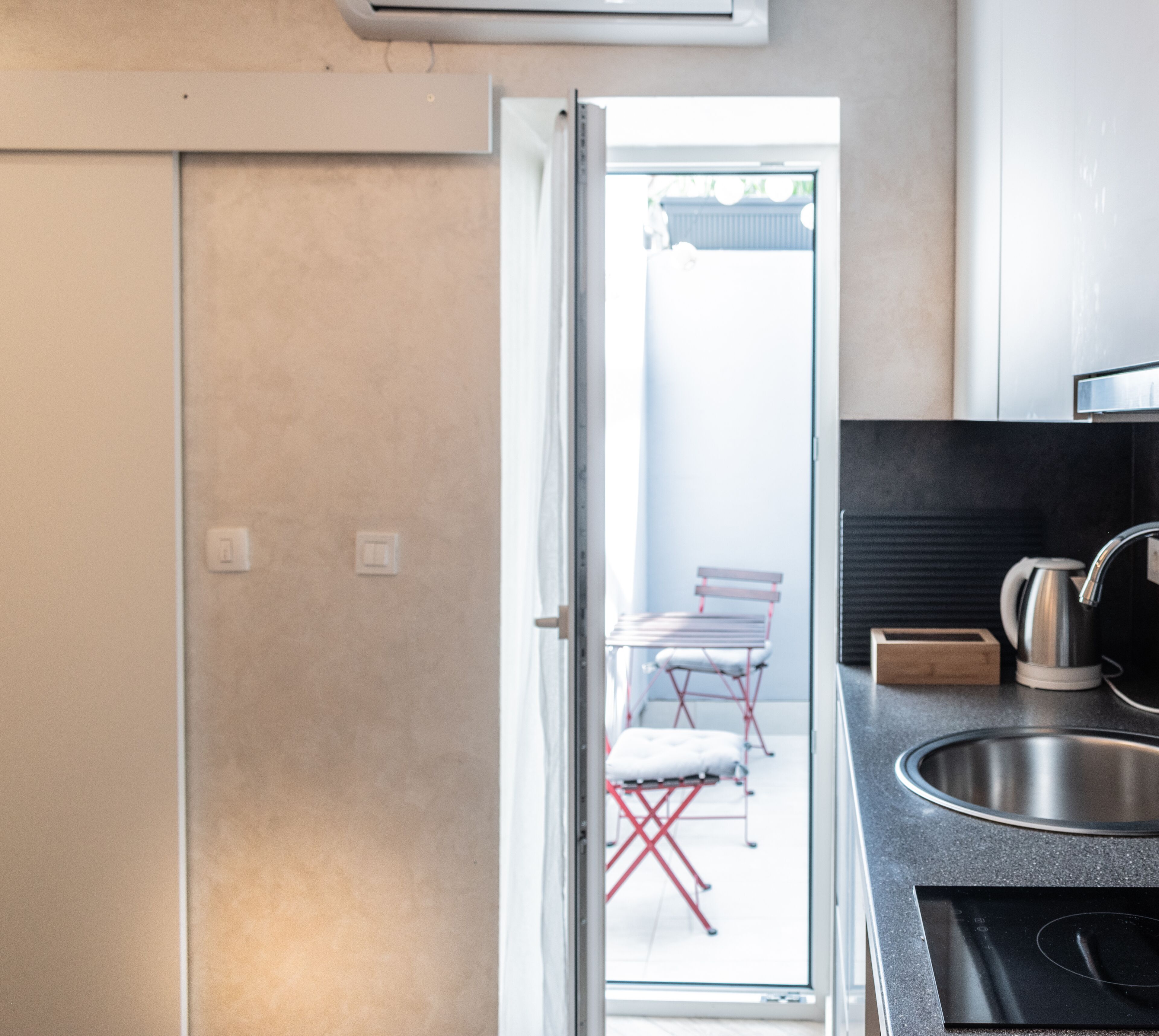 A modern kitchen with a stainless steel sink, a kettle, and a stove. There is a sliding glass door leading to a balcony with a chair and a table.
