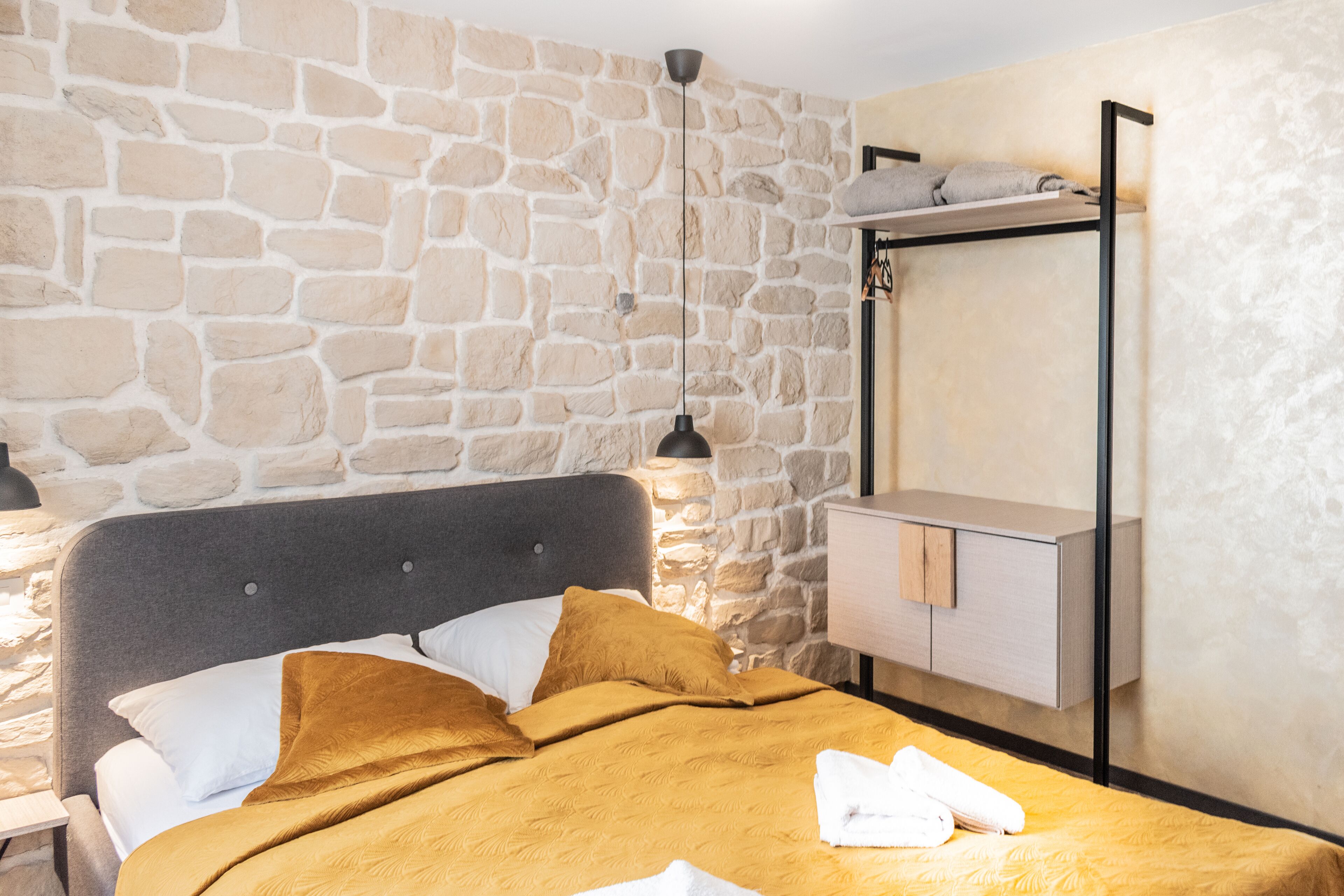 A room with a stone wall, a bed with a grey headboard, a bedside table with a lamp, and a wooden cabinet.