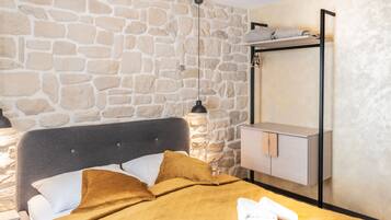 A room with a stone wall, a bed with a grey headboard, a bedside table with a lamp, and a wooden cabinet.