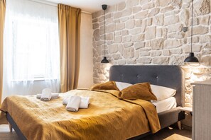 Una habitación de hotel con pared de piedra, una cama con cabecera, dos lámparas de noche y una ventana con cortinas.