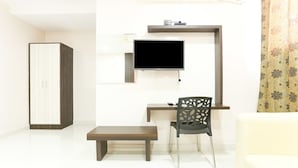 A modern living room with a flat-screen TV mounted on the wall, a wooden bench, a black chair with a unique cut-out design, and a white sofa. - Hotel Habitat Comforts (Bengaluru)