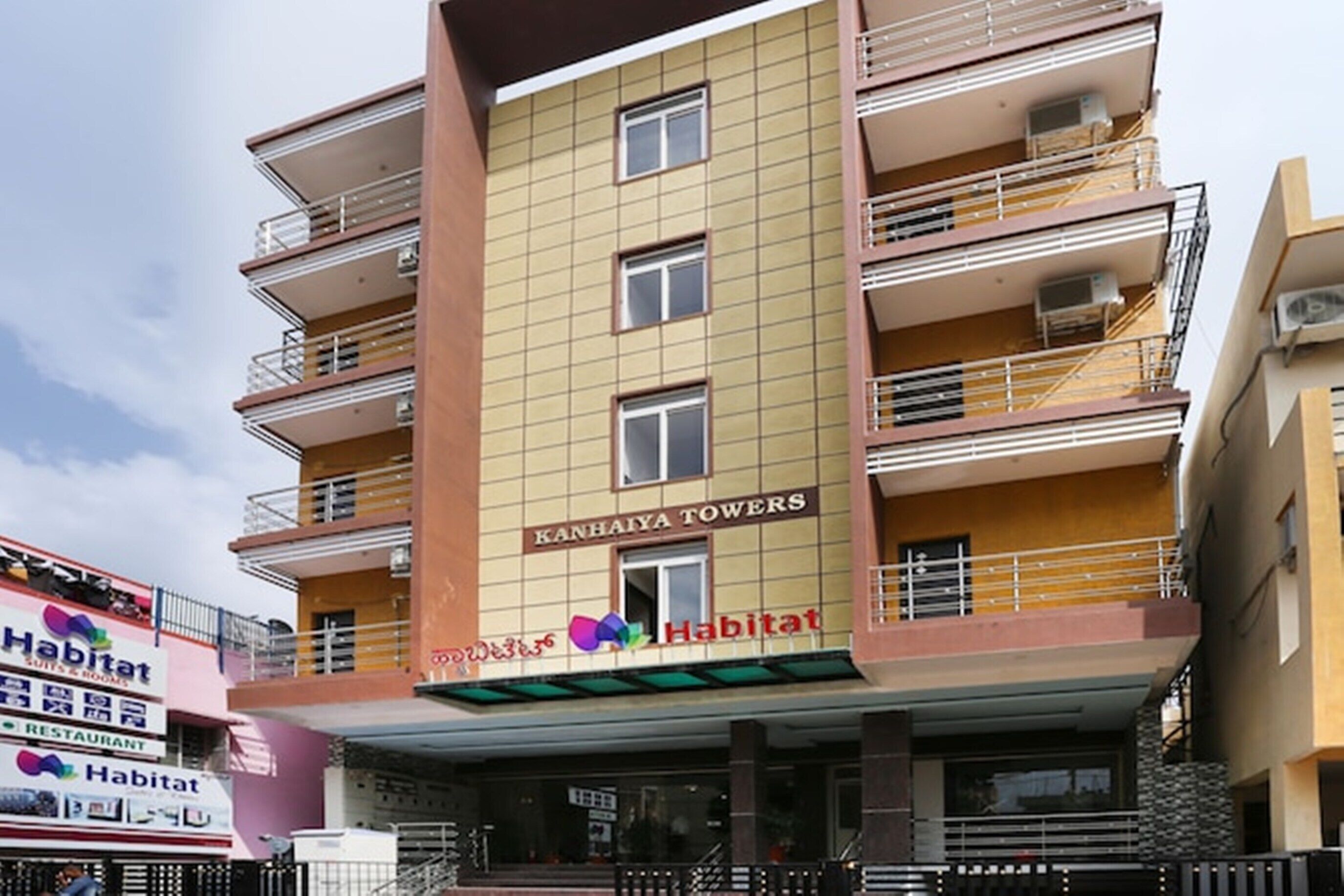 Un edificio de varios pisos con balcones y unidades de aire acondicionado. El edificio tiene un letrero que dice "Kanhaiya Towers" y "Habitat", indicando que es un hotel.