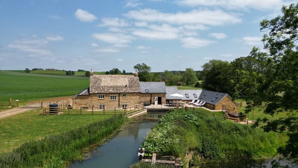 Une ferme en pierre avec une rivière en contrebas, une terrasse en bois et un champ verdoyant.