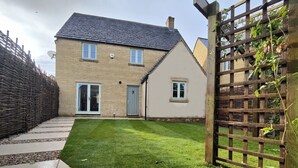 A two-story house with a sloped roof, a front door, and a small front yard with a wooden fence and a trellis. - Three bedroom dog friendly holiday cottage in the Cotswolds - Cedar Cottage (Bourton-on-the-Water)