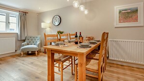 A dining area with a wooden table set for four, surrounded by wicker chairs. A clock and a framed picture adorn the walls. - Three bedroom dog friendly holiday cottage in the Cotswolds - Cedar Cottage (Bourton-on-the-Water)