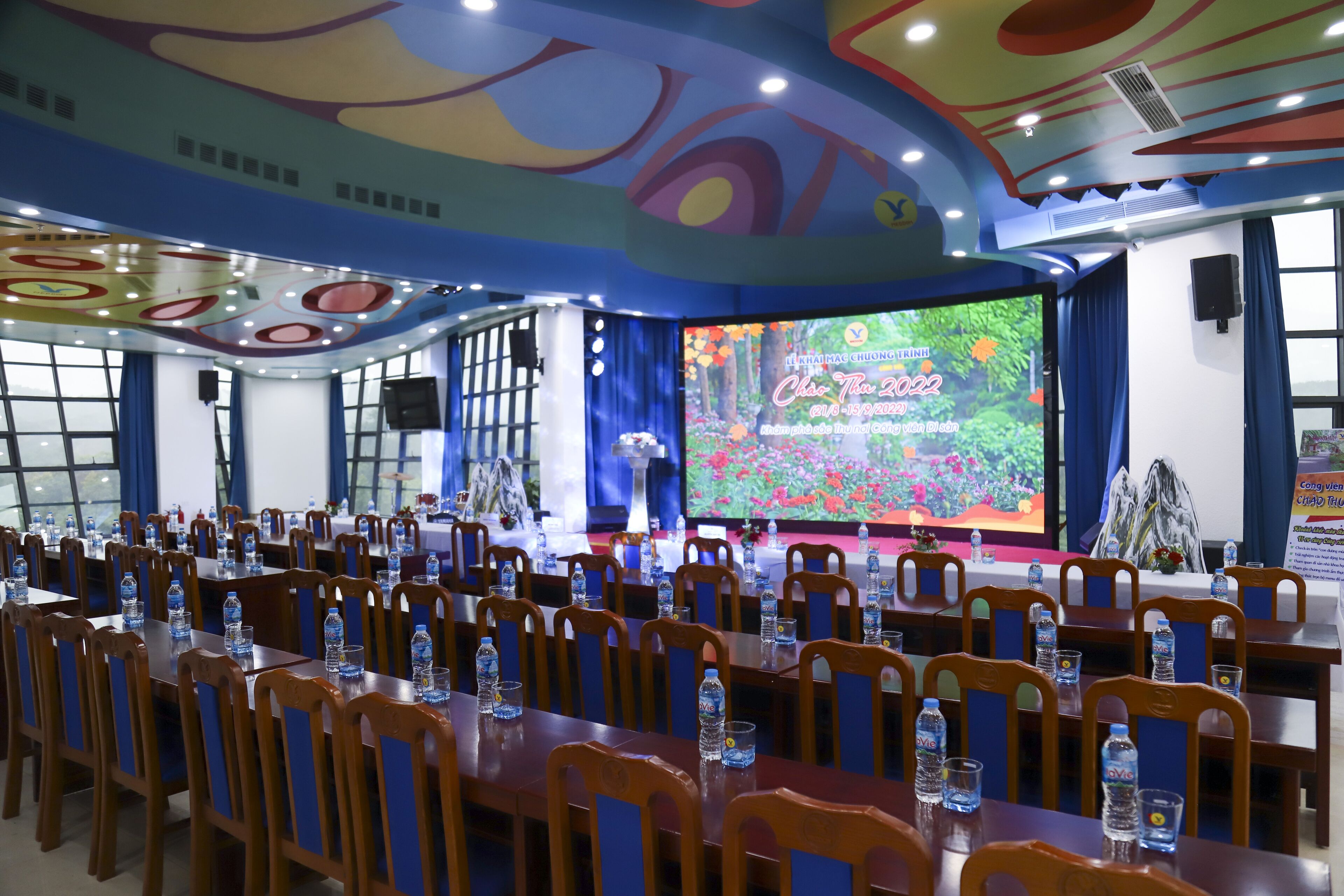 A conference room with rows of wooden tables and chairs, a stage with a large screen, and a colorful ceiling design.