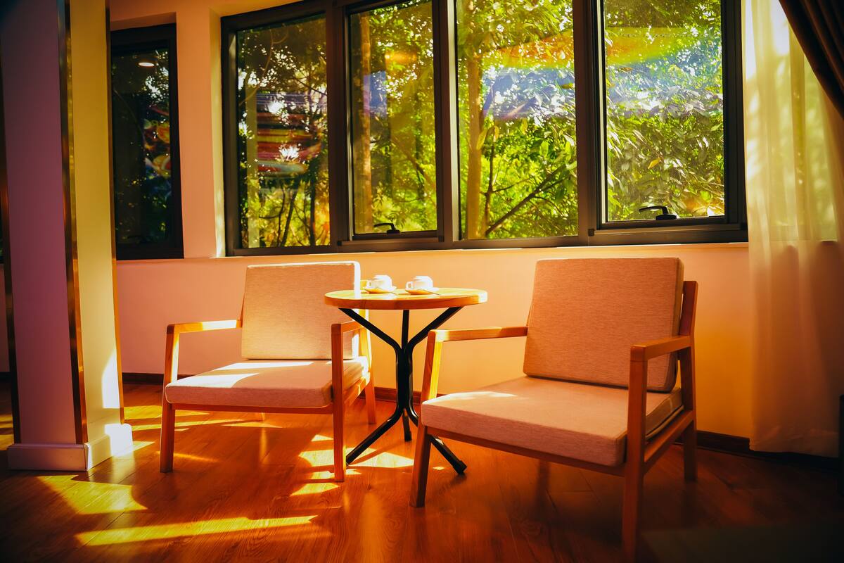 A room with two wooden chairs, a small round table, and large windows with a view of greenery outside.