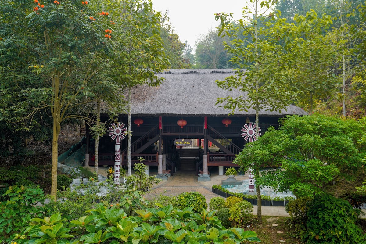 A traditional thatched-roof building with a staircase leading to a covered entrance, surrounded by lush greenery and a decorative windmill.