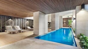 Uma área interna de piscina moderna, com espreguiçadeiras, teto de madeira e piscina azul cristalina.