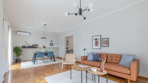 A modern living room with a wooden floor, a sofa, a coffee table, and a dining area with chairs and a table. - Homey 2BR Apartment in Cholargos (Cholargos)