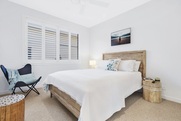 Een slaapkamer met een bed, een stoel, een ronde houten tafel, een nachtkastje en een raam met zonwering.