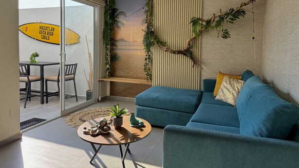 A modern living room with a blue sofa, a round coffee table with decorative items, a glass door leading to a balcony with a surfboard, and a wall-mounted plant. - 5 minutes from the beach/private pool by Gpsrentas (Mazatlán)