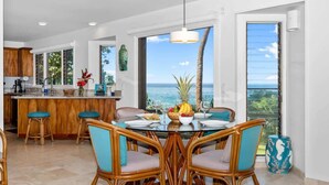 A modern kitchen with a central island, a dining area with wicker chairs, and a view of the ocean. - Oceanfront Luxury House, Pool & AC, Near Magic Sands (Kailua-Kona)