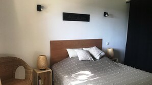 A bedroom with a wooden headboard, a bed with patterned bedding, a wicker chair, a small side table with a lamp, and a wall-mounted sign.