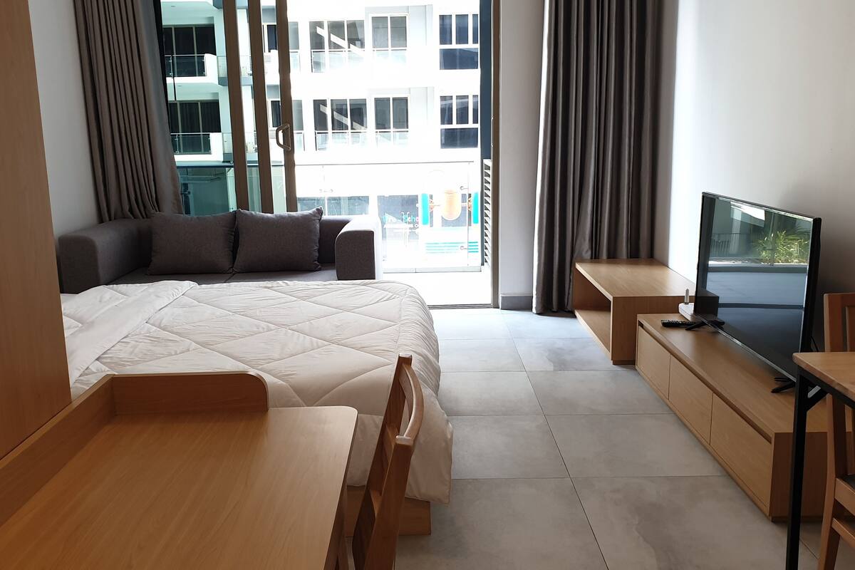 A modern hotel room with a bed, a desk, a chair, a TV, and a view of the city through large windows.