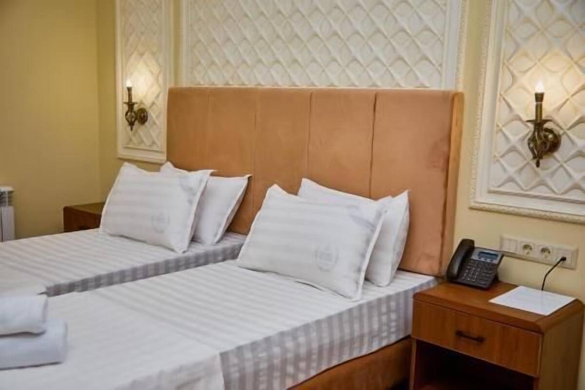 A hotel room with two beds, a wooden headboard, a bedside table with a phone, and a wall-mounted light fixture.