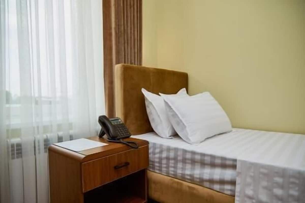 A hotel room with a wooden nightstand, a telephone, a bed with white pillows, and a window with sheer curtains.