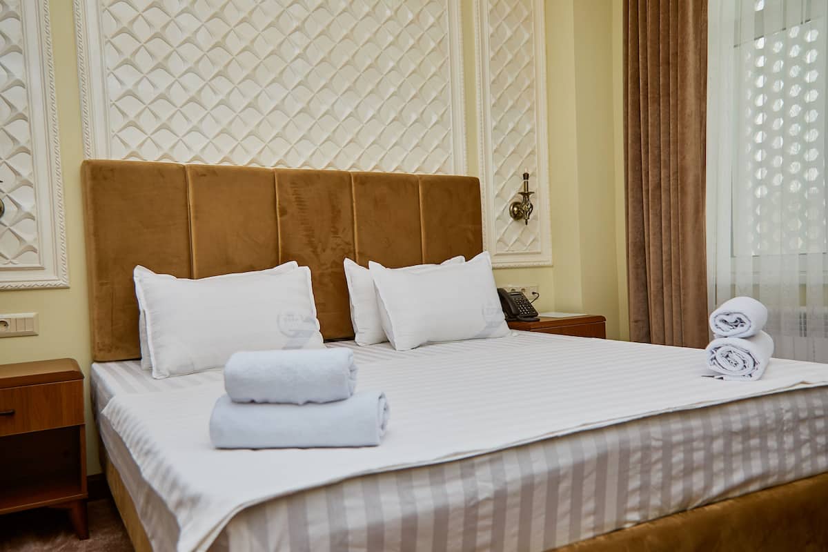 A neatly made bed with white linens and two rolled towels, a wooden nightstand with a phone, and a decorative wall panel.