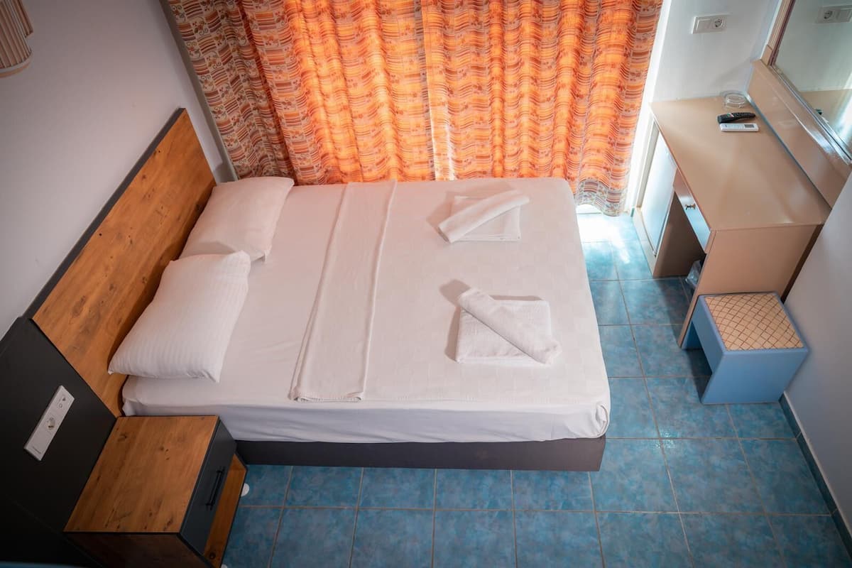 A neatly made bed with white linens and towels, a wooden headboard, a small bedside table with a remote control, and a patterned curtain.