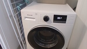 Lavadora blanca de carga frontal, compacta, con pantalla digital, ubicada en un lavadero pequeño que incluye un tendedero de metal y una cesta encima.