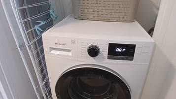 Une machine à laver compacte, blanche, à chargement frontal, dotée d’un écran numérique, installée dans une petite buanderie avec un étendoir métallique et un panier placé sur le dessus.