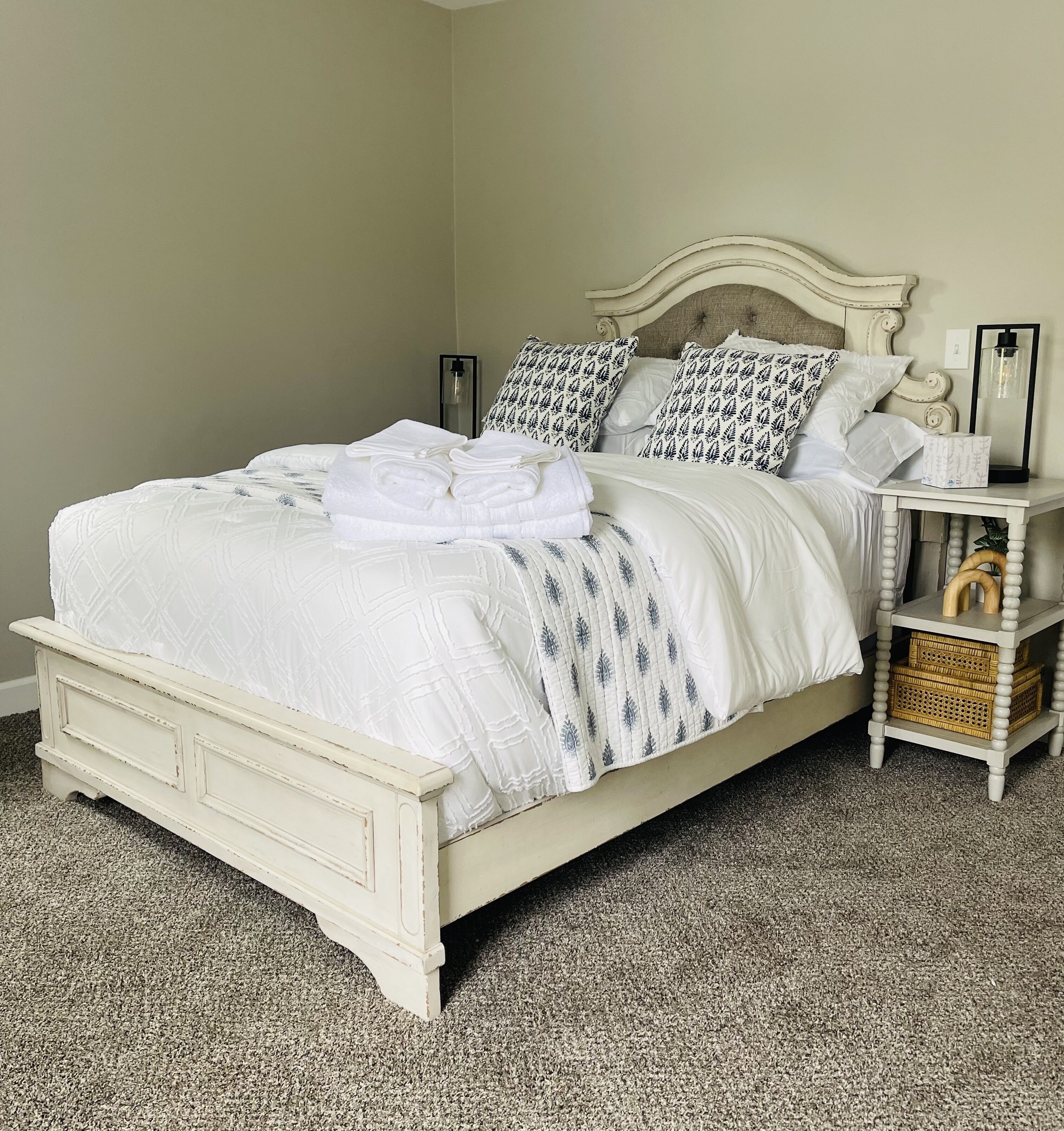 A neatly made bed with white linens and patterned pillows, a bedside table with a lamp, and a small wooden side table with baskets.