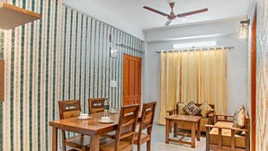 A dining area with a wooden table and chairs, a sofa set, a ceiling fan, and a window with curtains. - THE KASAULI TRAIL (Kasauli)