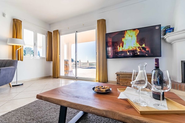 Ein modernes Wohnzimmer mit Fernseher, der einen Kamin zeigt, einem Esstisch mit Weingläsern und einer Flasche sowie Blick durch Glastüren.