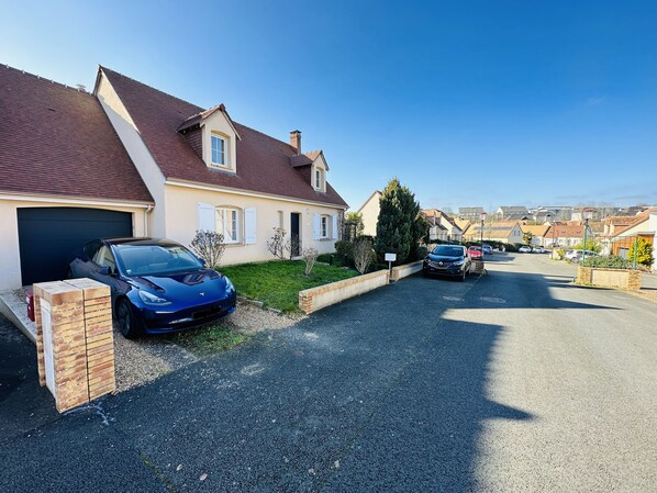 A residential street with houses, a blue car parked in front of one house, and another car parked further down the street.