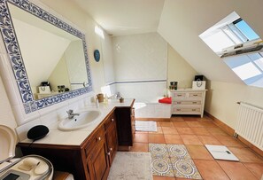 A bathroom with a large mirror, white tiles, a wooden vanity, a bathtub, and a skylight.