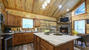 A wooden cabin interior with a kitchen, dining area, and fireplace. - Moonlit Oaks Retreat | Romantic Broken Bow Cabin with Hot Tub, Fire Pit & Fireplace (Broken Bow)