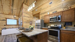 A wooden cabin interior with a kitchen featuring a large island, stainless steel appliances, and wooden cabinets. - Moonlit Oaks Retreat | Romantic Broken Bow Cabin with Hot Tub, Fire Pit & Fireplace (Broken Bow)