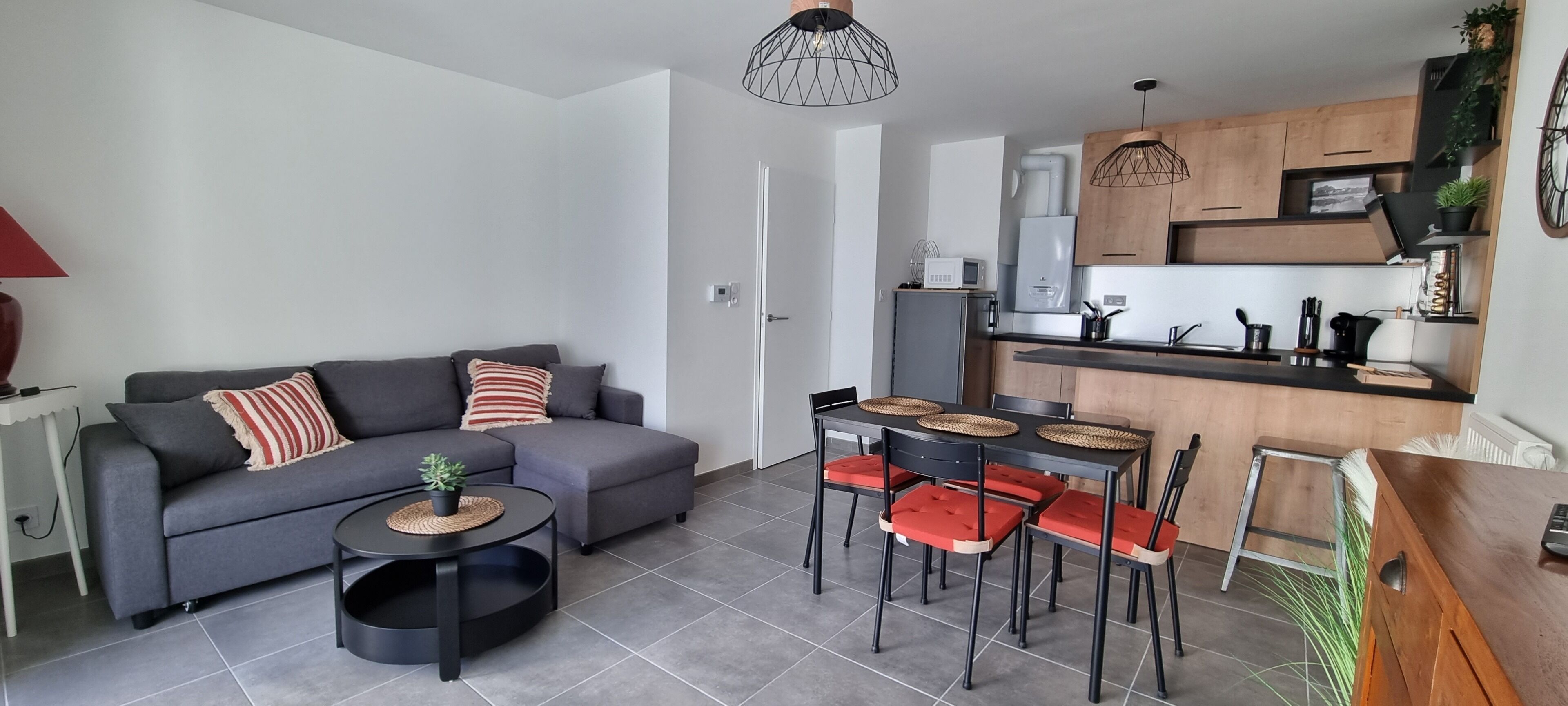 Een moderne woonkamer met een grijze bank, rode en witte gestreepte kussens, een klein rond tafeltje met een plant, en een eethoek met een houten tafel en rode stoelen.
