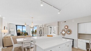 A modern kitchen with white cabinetry, a central island, and a dining area with white chairs and a table. - Captains Quarters 3BR Villa Steps from the Ocean (Pawleys Island)