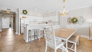 A modern kitchen with white cabinetry, a central island, and a dining area with white chairs and a wooden table. - Captains Quarters 3BR Villa Steps from the Ocean (Pawleys Island)