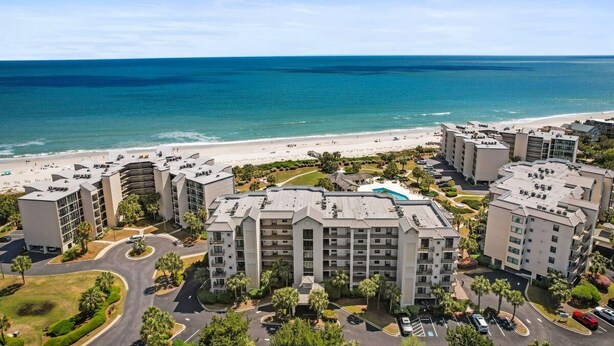 A beachfront hotel with multiple buildings, a parking area, and a beach in the background. - Captains Quarters 3BR Villa Steps from the Ocean (Pawleys Island)