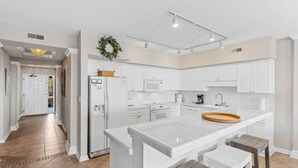 A modern kitchen with white cabinetry, a central island with stools, and a view into the adjacent dining area. - Captains Quarters 3BR Villa Steps from the Ocean (Pawleys Island)