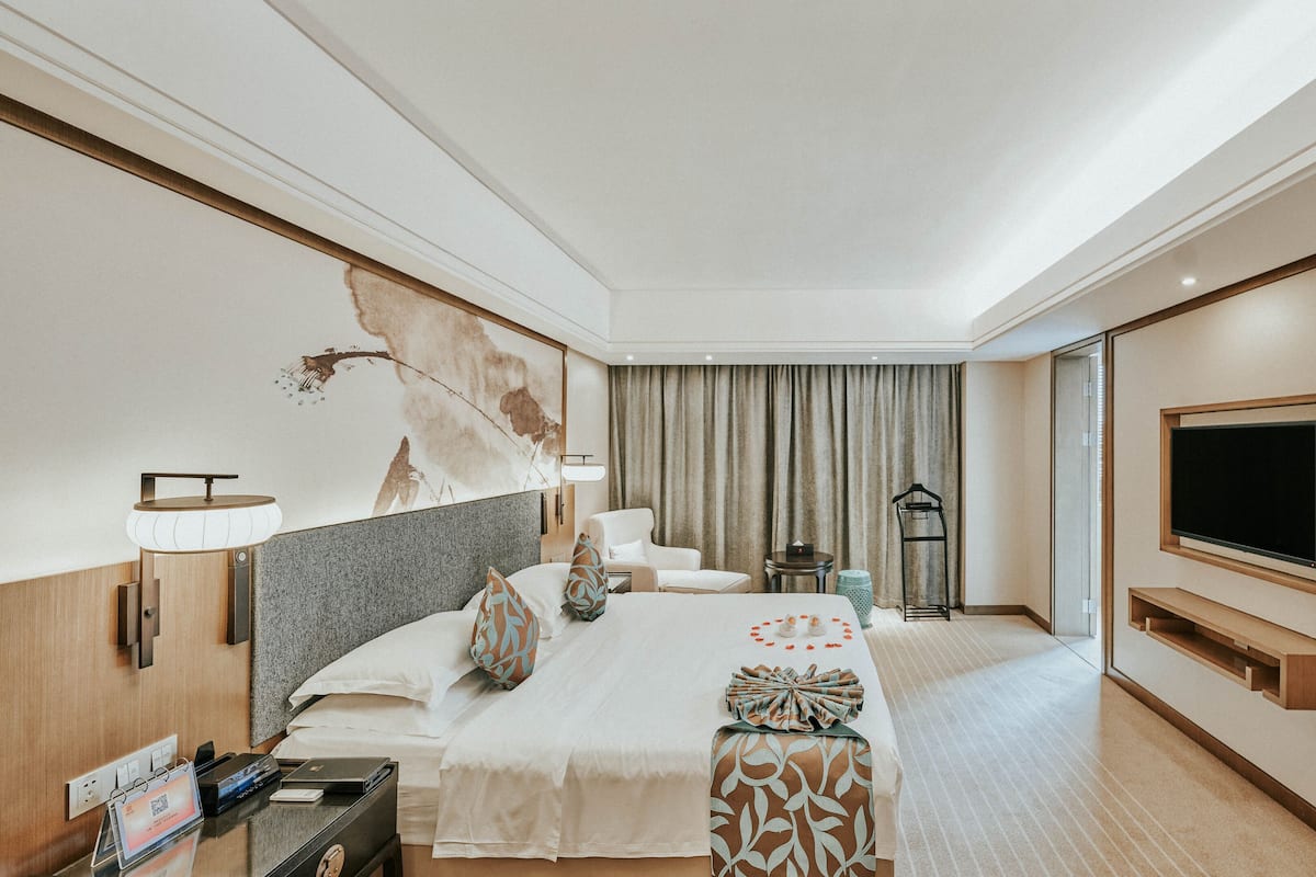 Ein Hotelzimmer mit Bett, Nachttisch, Lampe, Fernseher und einem großen Fenster mit Vorhängen.