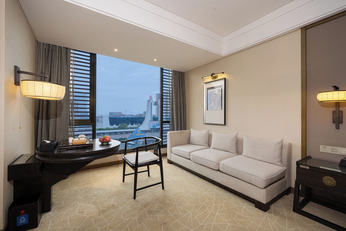 Ein modernes Hotelzimmer mit einer Couch, einem kleinen Tisch und einem Stuhl. Ein großes Fenster bietet Blick auf die Stadtlandschaft.