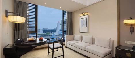 A modern hotel room with a sofa, a small table, and a chair. There is a large window with a view of the cityscape.