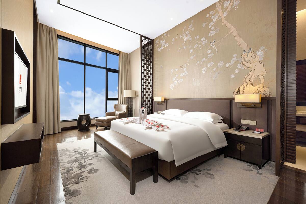 Ein modernes Hotelzimmer mit einem großen Bett, einem Schreibtisch, einem Sessel und einem großen Fenster mit Blick zum Himmel.